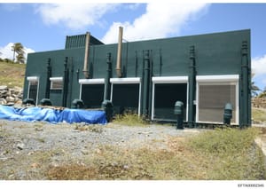 This photograph shows a utilitarian green metal building with multiple roll-down doors, ventilation pipes, and cooling systems. The structure appears to be industrial or infrastructure-related, situated on a hillside with palm trees visible in the ba