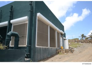 This is a photograph of an industrial or utility building featuring a dark teal/green exterior wall with white-trimmed louvered windows, metal piping, and a gravel/stone foundation. The structure appears to be located in a tropical or semi-arid locat