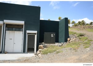 This is an evidence photograph showing the exterior of a modern dark-colored building or structure with multiple entrances and utility equipment. The building features white-trimmed doors and a dark metal utility box, situated on a rocky/gravel terra