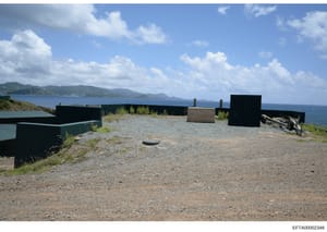 This is a photograph of a waterfront property featuring multiple black-painted structures and storage buildings on a gravel compound overlooking a blue ocean with mountainous islands visible in the distance. The image appears to be evidence documenta
