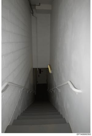 This is a photograph of a narrow, institutional-style stairwell with white cinderblock walls, concrete steps, and minimal lighting. The space appears to be a basement passage or utility area, showing metal handrails and basic infrastructure. The imag