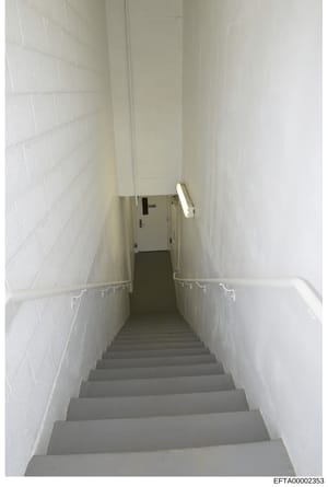 This is a photograph of a narrow interior stairwell with white subway tile walls, gray concrete steps, and a metal handrail. The stairwell appears to be in a basement or lower level of a building, with fluorescent lighting visible at the top. The ima