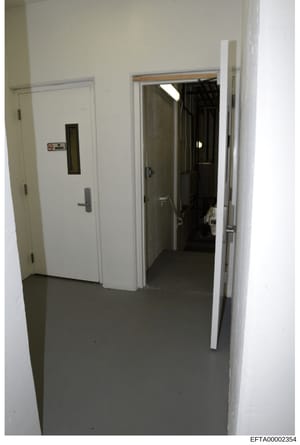 This photograph shows an interior hallway or corridor with white walls and a dark concrete floor. The image features an elevator on the left side marked with a sign, and open doorways leading to other rooms or spaces. The photo appears to be evidence