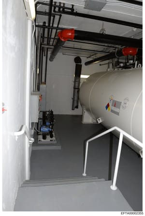 This is a photograph of an interior utility or mechanical room, likely from a residential or commercial property, showing a large cylindrical tank system with piping infrastructure, red safety equipment, and control mechanisms. The image appears to b