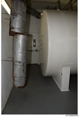 This photograph shows an interior basement or utility room space containing a large white cylindrical storage tank (likely propane or fuel storage) and a heavily corroded metal pipe running vertically along the left wall. The image appears to be evid