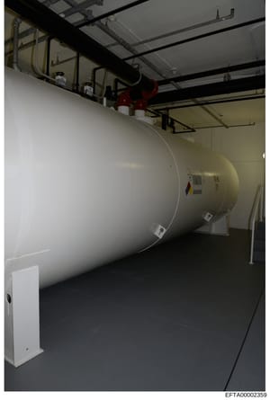 This is a photograph of an industrial storage tank or pressure vessel installed in what appears to be a basement or utility room. The tank is cylindrical, cream/beige colored, with overhead piping and mechanical systems visible. The image shows infra