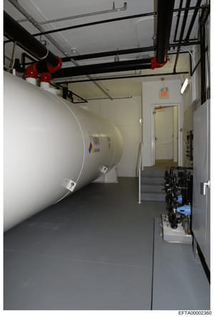 This photograph shows an interior mechanical or utility room, likely part of a residential or commercial property. The image depicts a large white cylindrical tank (possibly for water, heating, or storage), overhead piping infrastructure with red iso