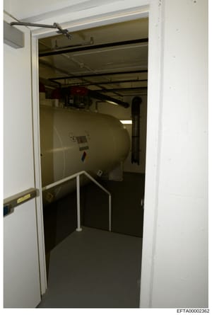 This is an evidence photograph of a mechanical or utility room featuring a large cylindrical storage tank, exposed ceiling beams, metal handrails, and various equipment. The image appears to be documentation of property infrastructure, likely taken d
