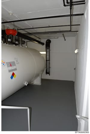 This is an evidence photograph showing the interior of a utility or mechanical room, featuring a large cylindrical heating/storage tank with hazard labeling, black support beams, electrical conduits, and a red emergency device mounted on the ceiling.
