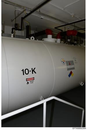 This is a photograph of a large industrial diesel fuel storage tank labeled '10-K' with a 'DANGER - DIESEL FUEL' warning label. The tank appears to be located in a basement or underground facility and is equipped with piping, valves, and safety equip