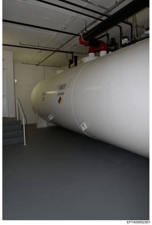 This photograph shows a large cylindrical storage tank installed in an interior basement or utility room space. The tank appears to be a commercial-grade heating oil, water, or propane storage vessel with visible control equipment and safety features