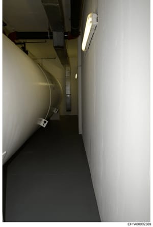 This is an evidence photograph showing a narrow interior hallway or corridor with white walls, fluorescent lighting, and visible mechanical infrastructure including pipes and conduit work. The image appears to be documentation of a building interior,
