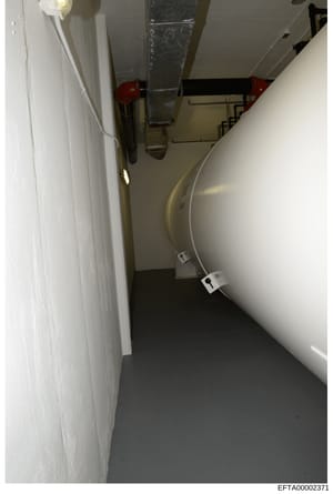 This is an evidence photograph documenting a basement or utility room interior, showing HVAC/mechanical systems including ductwork, piping, and what appears to be a large cylindrical tank or equipment. The space has white-painted walls, dark flooring