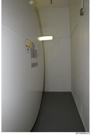 This is a photograph of a narrow interior hallway or corridor with white painted walls, a dark floor, and fluorescent ceiling lighting. The space contains two visible posters or signs on the wall, including what appears to be a Pittsburgh Steelers lo