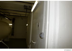 This photograph shows an interior room or utility space, likely part of a residential or commercial property. The space appears largely empty with exposed ceiling beams, fluorescent lighting, and various mechanical/electrical fixtures. The image may 