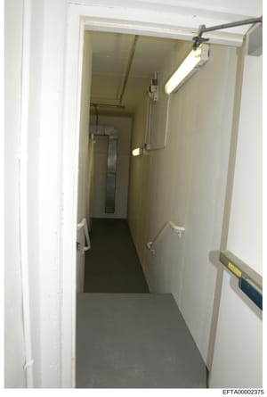 This is a documentary photograph of an interior hallway or corridor, likely taken during an investigation of one of Jeffrey Epstein's properties. The image shows a narrow, institutional-style passageway with beige/cream colored walls, fluorescent lig