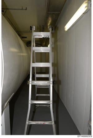This is an evidence photograph showing the interior of a narrow hallway or corridor in what appears to be a basement or utility area, with an aluminum extension ladder positioned in the center. The space features plain white walls, fluorescent lighti