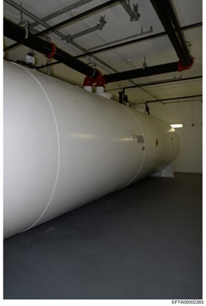 This is a photograph of a large cylindrical white storage tank or industrial vessel mounted horizontally in an indoor warehouse or industrial facility. The tank is suspended by red and black overhead support structures and piping systems. The image a