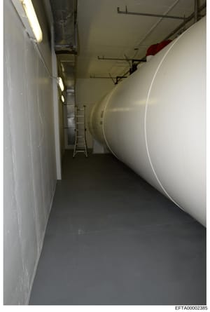 This photograph shows a narrow basement or utility room corridor with a large white cylindrical storage tank running along the right side. The space includes fluorescent lighting, exposed piping, a metal ladder, and what appears to be mechanical infr