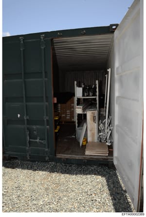 This photograph shows the interior of a large shipping container or storage unit with double doors open, revealing various items stored inside including what appears to be furniture, boxes, wine bottles, and other property. The container shows eviden