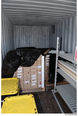 This photograph shows the interior of a shipping container or storage unit containing stacked wooden pallets with labeled packages covered in black plastic sheeting, along with yellow plastic bins and white metal shelving. The image appears to be evi