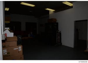 This is a photograph of an interior commercial or warehouse-type space showing a large empty room with fluorescent ceiling lights, white walls, and dark flooring. The room contains several cardboard boxes (including one labeled with 'Fiber' branding)