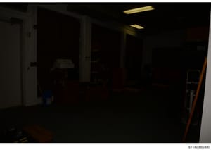 This photograph shows the interior of a dark, sparsely furnished room, likely part of a property being documented during the investigation. The image captures what appears to be a utility or storage area with minimal visible furnishings and equipment