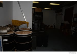 This is an evidence photograph showing the interior of what appears to be a storage or utility room. The image depicts large black drums or containers with lids, wooden furniture including a chair, metal equipment including what appears to be a metal