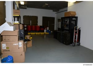 This is a photograph of an industrial or commercial storage space containing various equipment, cardboard boxes, and materials. The room features a Siglent device, red drums, black equipment cabinets, a hand truck, and multiple packed boxes labeled w