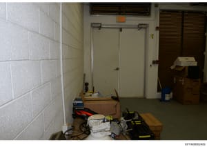 This is a photograph of an institutional or commercial storage room, showing white brick walls, fluorescent lighting, closed doors, and various stored items including electrical equipment, boxes, and tools. The image appears to be from a property sea