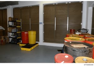 This photograph shows an interior storage area containing multiple industrial drums (red and yellow barrels), shelving units, and hazard containment equipment. A 'NOTICE' sign referencing waste oil is visible on the wall. The space appears to be a ba