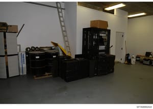 This is an evidence photograph depicting the interior of a storage or warehouse space containing numerous seized items including black storage containers, furniture, equipment, and various household items. The image appears to be part of the forensic