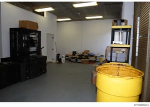 This photograph documents an interior space, likely a storage area or warehouse, containing various equipment, boxes, and materials. The image shows industrial shelving, a large yellow barrel or tank, black storage cabinets, and numerous cardboard bo