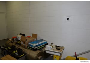 This is an evidence photograph of a storage or utility room containing various equipment and items including pumps, boxes, tools, and miscellaneous supplies. The room has white cinder block walls and concrete flooring. The letter 'D' is marked on the