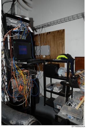 This is a photograph of a server room or technical equipment area showing networked computer hardware, networking equipment with extensive cabling, and storage infrastructure. The image appears to be evidence documentation from a property search, sho