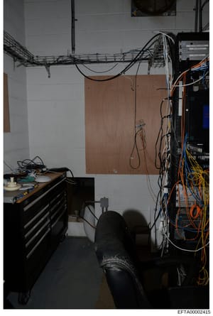 This is a photograph of an interior space containing computer/networking equipment, including a wall-mounted cable tray system, server or electronic devices on the right side, a wooden panel on the wall, and various cables and wiring. The image appea
