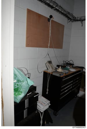 This photograph shows a basement or institutional room with white cinder block walls containing electronic equipment mounted on a wooden panel. The setup includes what appears to be surveillance or monitoring devices with cables, housed equipment on 