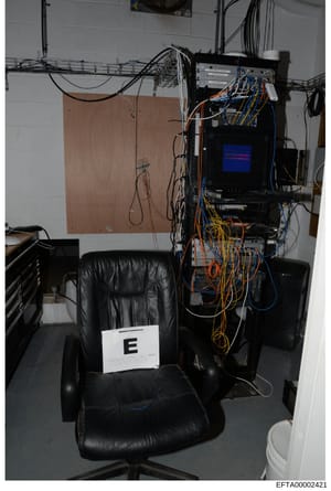 This is a forensic evidence photograph showing a black leather office chair marked with evidence label 'E', positioned in front of computer equipment with extensive orange and blue cabling, a monitor displaying a blue screen, and cable management inf