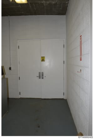 This is an evidence photograph showing an institutional or commercial interior hallway with white subway tile walls, a concrete floor, and a set of double white doors with electronic locks. A yellow 'Danger' warning sign is visible on the doors, and 