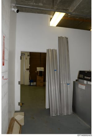 This photograph shows an interior room or storage area within a property under investigation, featuring industrial-style construction with white walls, concrete flooring, exposed ceiling infrastructure, and stored materials including metal panels and