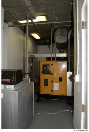 This photograph shows an interior utility or mechanical room containing a yellow Caterpillar diesel generator unit, fluorescent lighting fixtures, industrial piping, and various equipment boxes. The space appears to be a basement or utility area of a