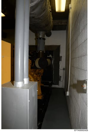 This is an evidence photograph showing the interior of a narrow mechanical or utility room containing industrial equipment. The image displays a large yellow and black generator or similar mechanical apparatus, a gray support column, concrete block w