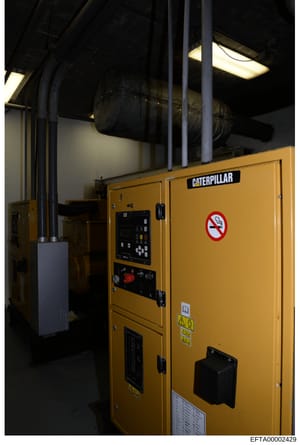 This is a photograph of a large yellow Caterpillar diesel generator unit installed in an industrial or commercial facility. The generator features control panels, warning labels, and no-smoking signage. The image documents equipment that may have bee