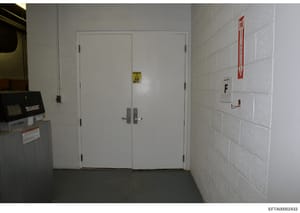 This is an evidence photograph showing an interior hallway or corridor with white-painted concrete block walls, institutional-style double doors with safety signage, and utility equipment. The image appears to be from a building inspection or evidenc
