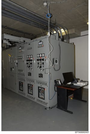 This is a photograph of an industrial electrical control room or equipment facility showing large gray electrical distribution panels, monitoring systems, control consoles, and overhead wiring infrastructure. The image appears to be documentation of 