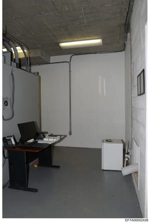 This is a photograph of a small interior room or office space, likely taken as part of the investigative documentation. The room contains basic office equipment including a desk with a computer monitor, phone, and other items, along with HVAC equipme