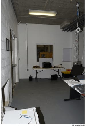 This is an evidence photograph showing the interior of a basement or utility room containing a folding work table, various tools and equipment, exposed ceiling infrastructure, and electrical wiring. The space appears to be used for storage or work pu