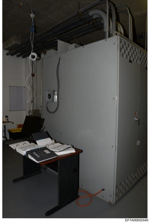 This photograph shows an interior laboratory or technical workspace containing a large white electrical/mechanical cabinet, a black work desk with documents and materials, overhead piping and electrical conduits, and various technical equipment. The 