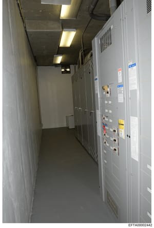 This is a photograph of a narrow, industrial corridor containing large electrical distribution panels and server equipment mounted on the right wall. The image shows rows of gray metal cabinets with circuit breakers, control panels, warning labels, a