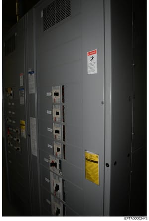 This photograph shows the interior of an electrical equipment room or utility closet, featuring a large gray panel cabinet with multiple circuit breakers or electrical control units, ventilation grilles, and prominent safety warning labels including 